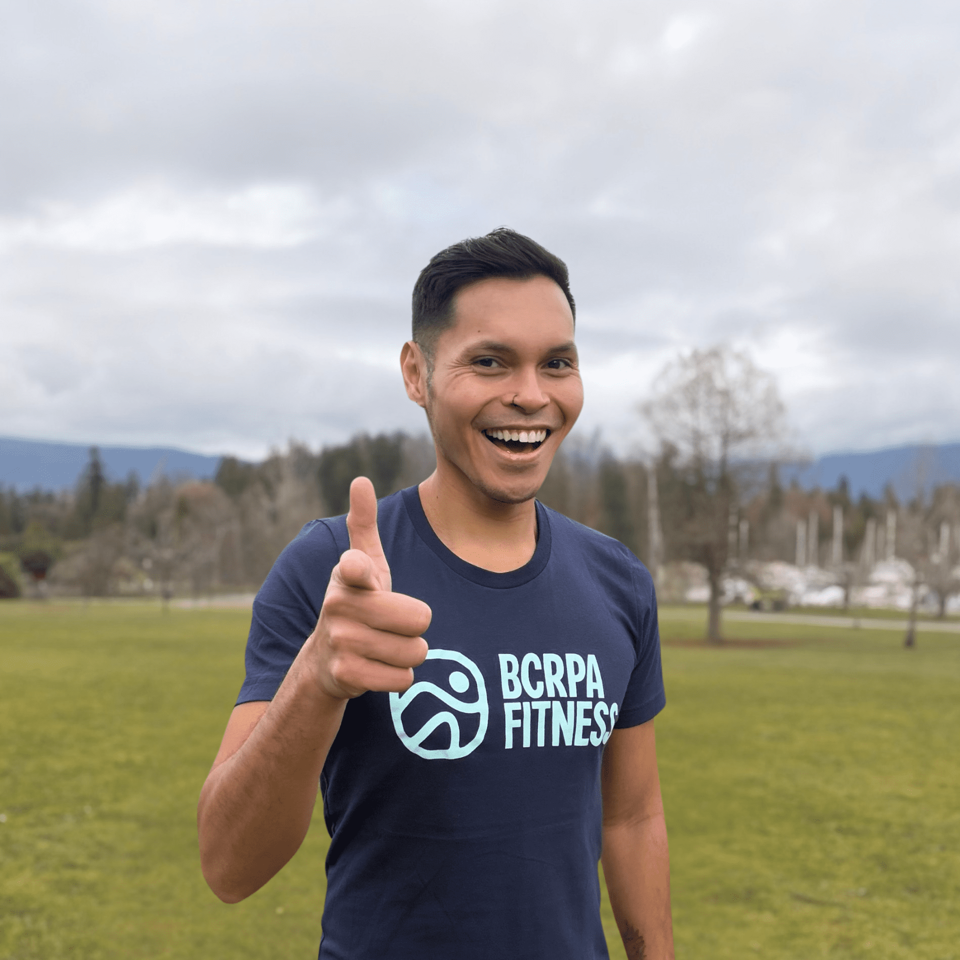 man wearing BCRPA Fitness t-shirt, smiling and pointing at the camera. Background is a grassy park with grey clouds in the sky.